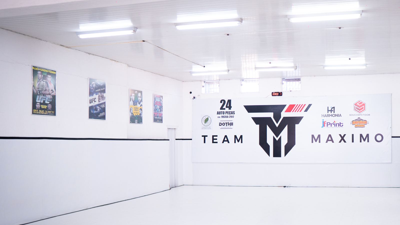 Tatame branco e amplo da academia, com a parede do logo Team Máximo ao fundo